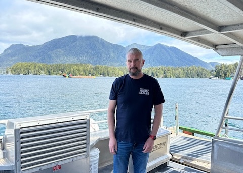 Ein Mann in einem T-Shirt mit der Aufschrift „West Coast Aquatic Safaris“, der auf dem Deck eines Bootes steht.
