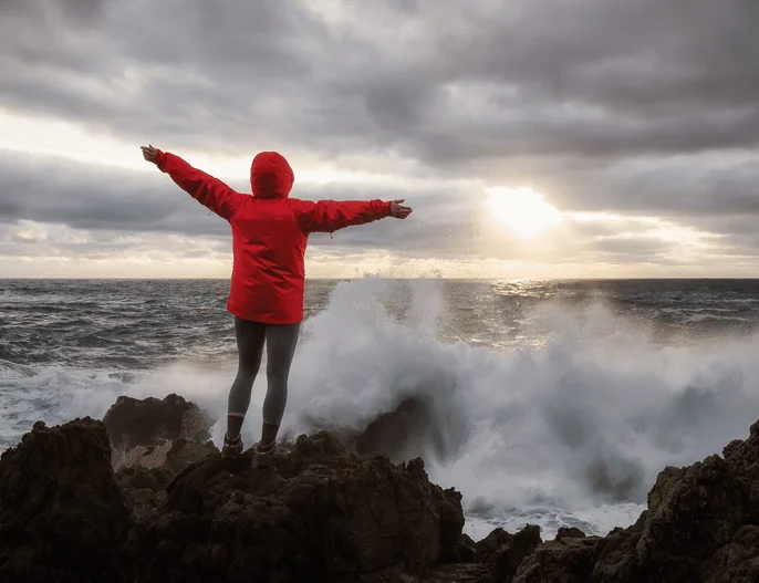 Person in a red jacket standing on coastal rocks as waves crash nearby.