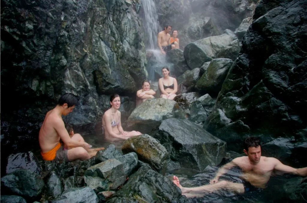 People relaxing in a natural hot spring surrounded by dark rocks and a waterfall.