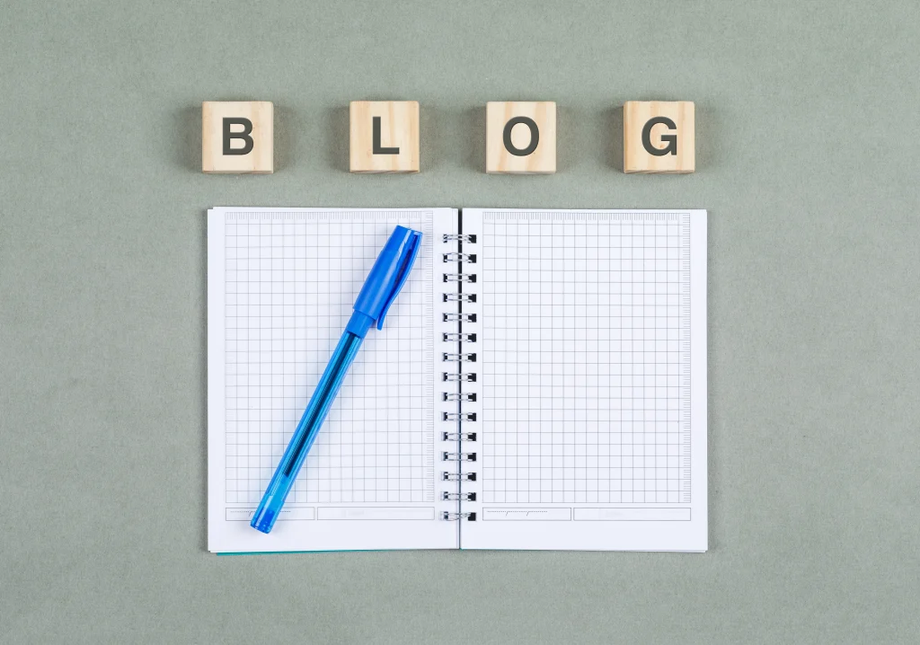 Open notebook and blue pen beneath wooden blocks spelling blog.